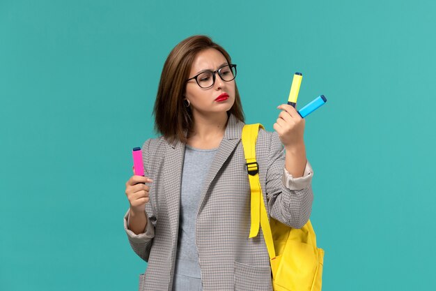 Front view of female student in grey jacket wearing her yellow backpack holding felt pens on the light blue wall