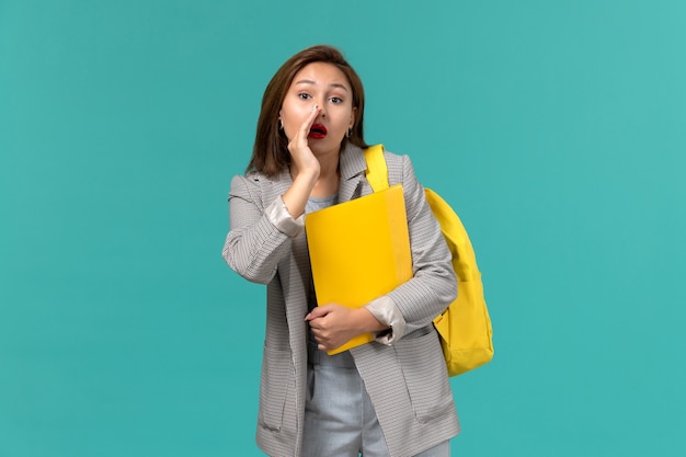 Front view of female student in grey jacket wearing her yellow backpack and holding files whispering on light blue wall