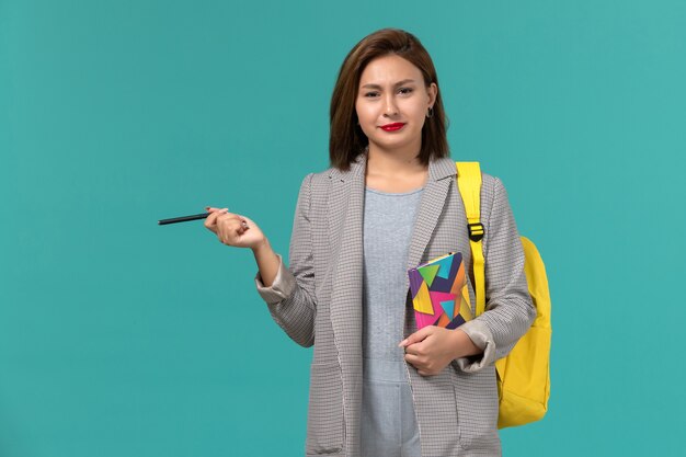 Front view of female student in grey jacket wearing yellow backpack holding copybook with pen on blue wall