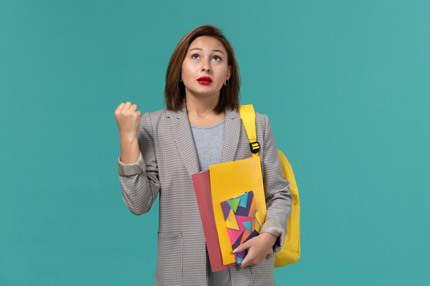 Front view of female student in grey jacket wearing yellow backpack holding files and copybook on blue wall