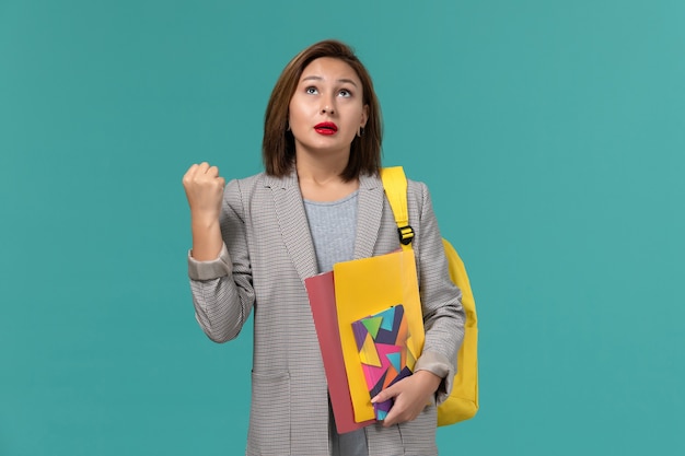 Front view of female student in grey jacket wearing yellow backpack holding files and copybook on blue wall