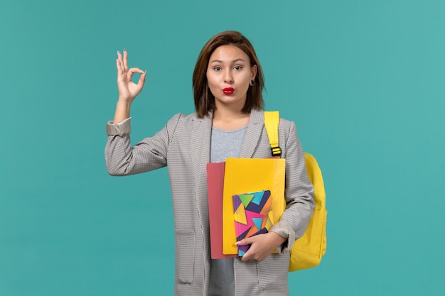 Front view of female student in grey jacket wearing yellow backpack holding files and copybook on blue wall