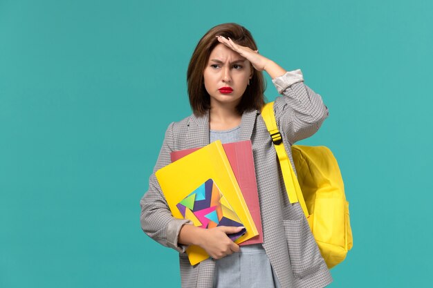 Front view of female student in grey jacket wearing yellow backpack holding files and copybook confused on blue wall