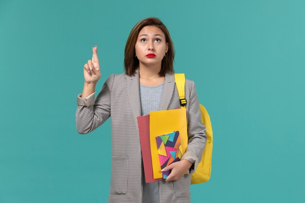 Front view of female student in grey jacket wearing yellow backpack holding files and copybook crossing her fingers on blue wall