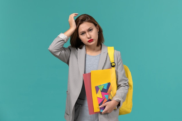 Front view of female student in grey jacket wearing yellow backpack holding files and copybook having headache on blue wall