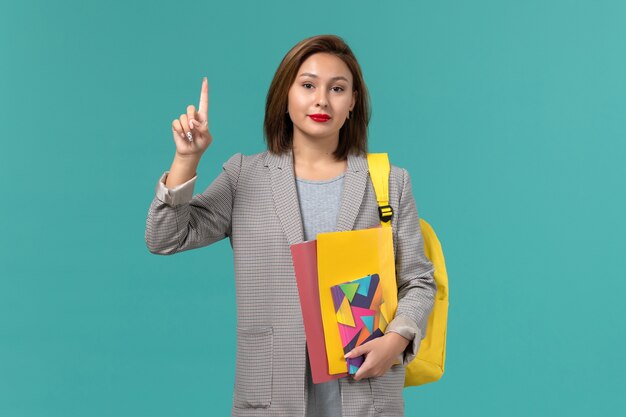 Front view of female student in grey jacket wearing yellow backpack holding files and copybook on the light blue wall