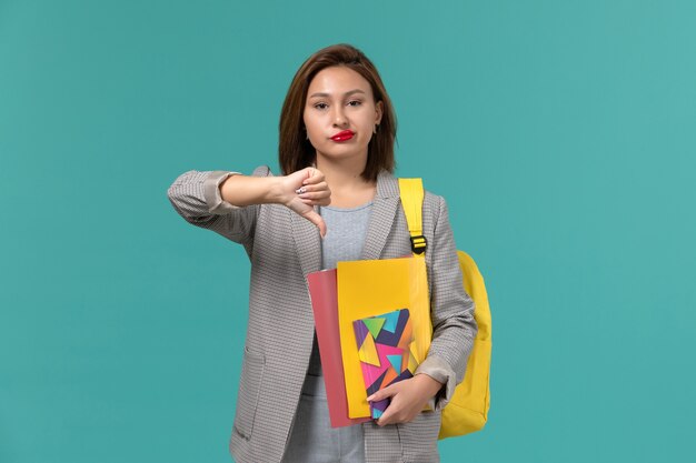 Front view of female student in grey jacket wearing yellow backpack holding files and copybook showing unlike sign on blue wall