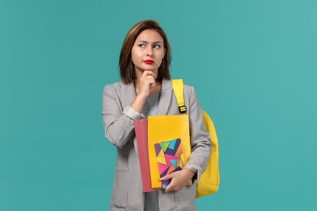 Front view of female student in grey jacket wearing yellow backpack holding files and copybook thinking on blue wall