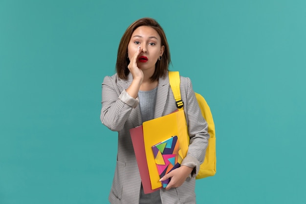 Front view of female student in grey jacket wearing yellow backpack holding files and copybook whispering on blue wall