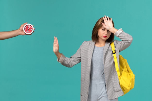 Front view of female student in grey jacket wearing yellow backpack on the light-blue wall