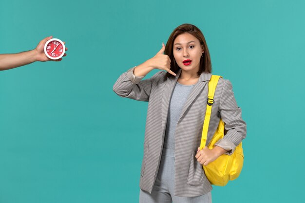 Front view of female student in grey jacket wearing yellow backpack on the light blue wall