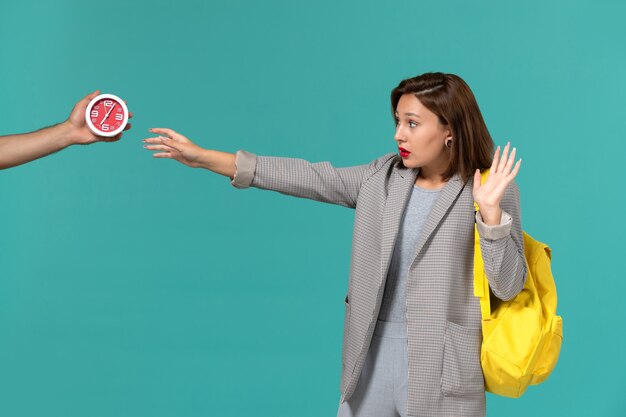 Front view of female student in grey jacket wearing yellow backpack looking at clocks on light-blue wall