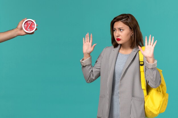 Front view of female student in grey jacket wearing yellow backpack looking at clocks on the light blue wall