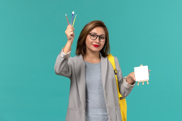 Front view of female student in grey jacket yellow backpack holding tassels and easel on the blue wall