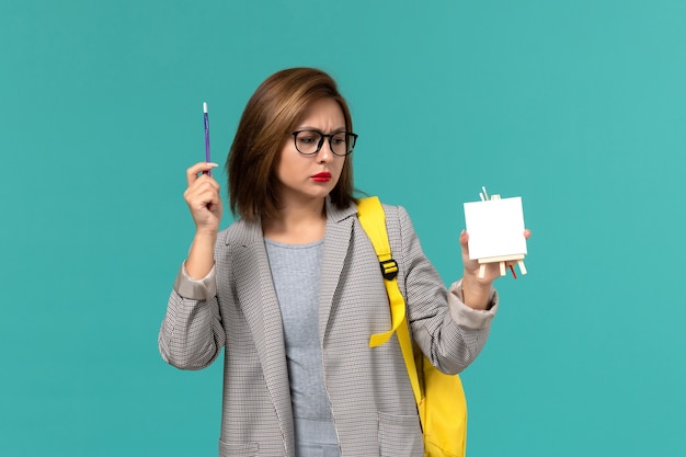 Front view of female student in grey jacket yellow backpack holding tassels and easel on light-blue wall