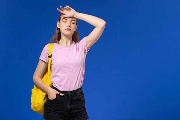 Front view of female student in pink t-shirt with yellow backpack on blue wall