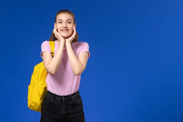 Front view of female student in pink t-shirt with yellow backpack smiling and posing on blue wall