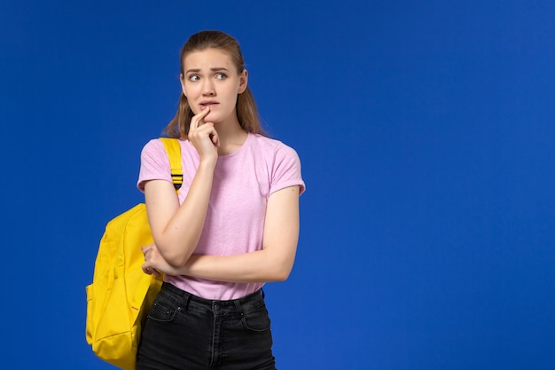 Front view of female student in pink t-shirt with yellow backpack and thinking on the blue wall