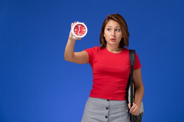 Front view of female student in red shirt with backpack holding clocks on the blue wall