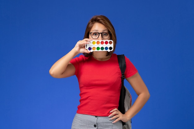 Free photo front view of female student in red shirt with backpack holding paints for drawing on blue wall