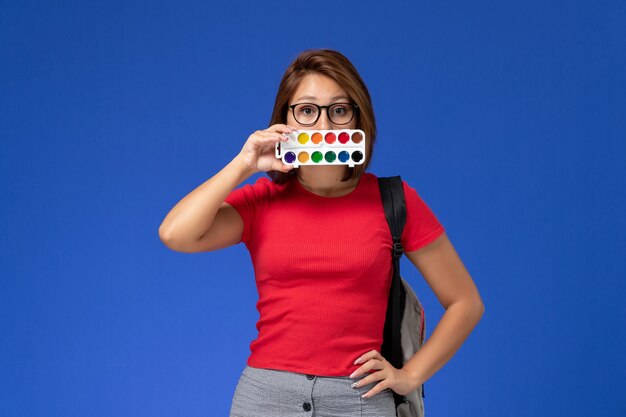 Front view of female student in red shirt with backpack holding paints for drawing on blue wall