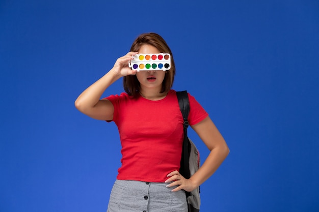 Free Photo front view of female student in red shirt with backpack holding paints for drawing on the blue wall