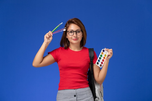 Front view of female student in red shirt with backpack holding paints for drawing and brushes on blue wall