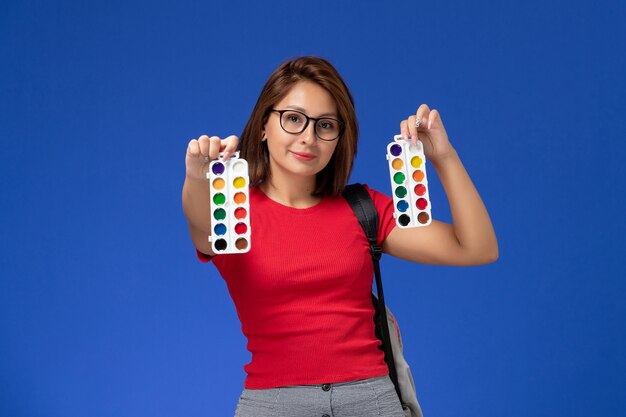 Front view of female student in red shirt with backpack holding paints for drawing smiling on blue wall