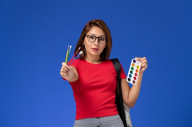 Front view of female student in red shirt with backpack holding paints for drawing and tassels on blue wall