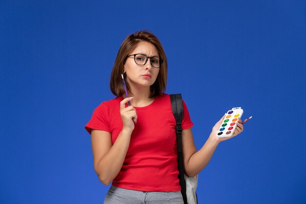 Front view of female student in red shirt with backpack holding paints for drawing and tassels on the blue wall