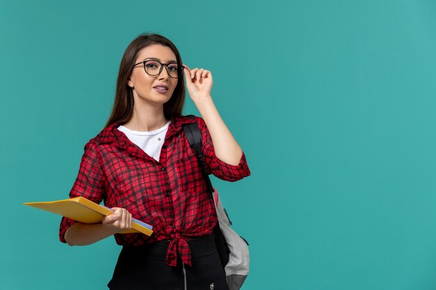 Front view of female student wearing backpack and holding files on light-blue wall