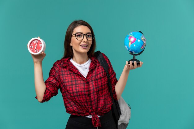 Front view of female student wearing backpack holding little globe and clocks on light blue wall