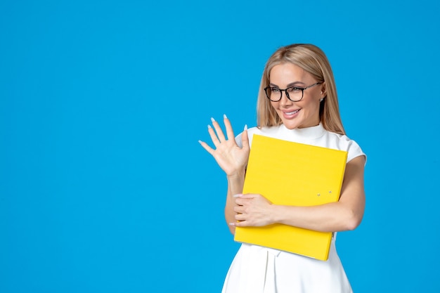 Front view of female worker in white dress holding folder and greeting someone on blue wall