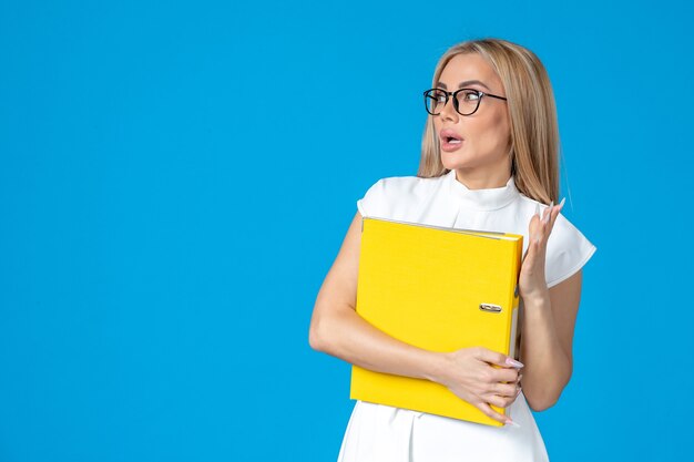 Front view of female worker in white dress holding yellow folder on blue wall