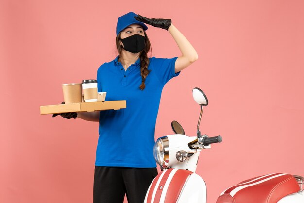 Front view of focused courier girl wearing medical mask gloves standing next to motorcycle holding coffee small cakes on pastel peach color background