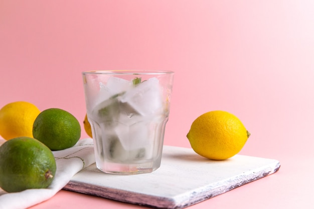 Front view of fresh cold lemonade with ice inside glass along with fresh lemons on the pink wall