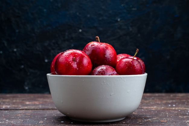 A front view fresh red plums mellow and ripe inside white plate on the wooden desk fruit pulp juice