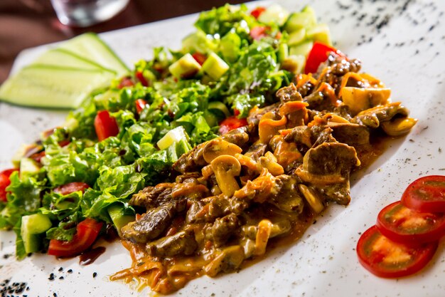 Front view fried meat with mushrooms in sauce with vegetable salad and slices of tomato and cucumber