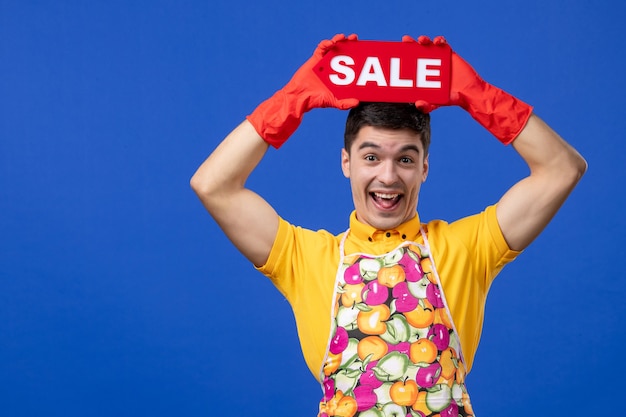 Front view funny cheerful housekeeper in yellow t-shirt raising sale sign over his head on blue space