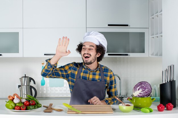 Front view of funny and emotional male chef with fresh vegetables and cooking with kitchen tools and saying hello in the white kitchen