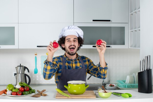 Front view of funny and emotional male chef with fresh vegetables holding red peppers in the white kitchen