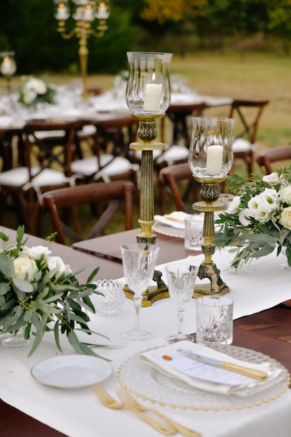 Front view of glassware and cutlery served on the wooden table with floral compositions and candlesticks outdoors