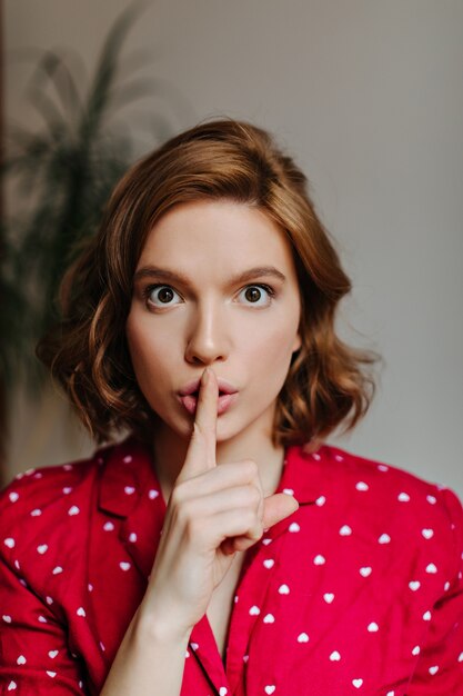 Front view of gorgeous woman showing hush sign. Indoor shot of curly female model in red pajama.