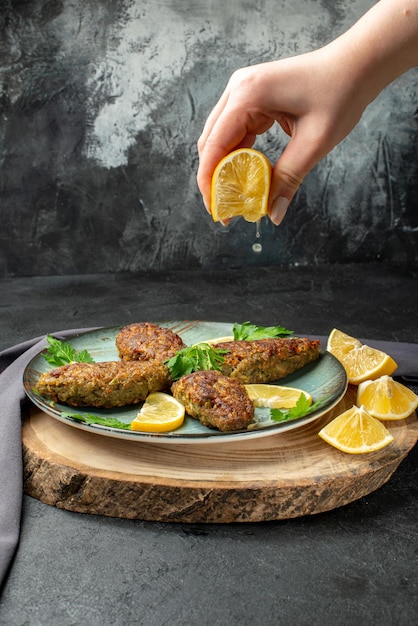 Front view of hand squeezing limon on delicious cutlets served with greens on wooden cutting board black towel on dark color background