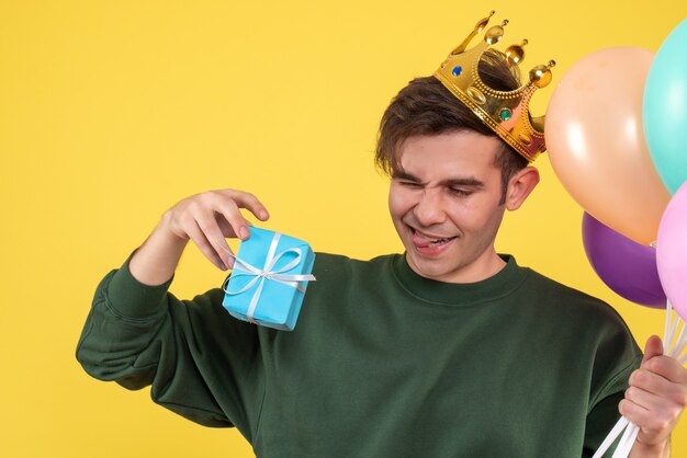 Front view handsome young man with crown holding balloons and gift on yellow 