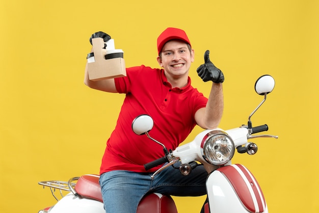 Front view of happy courier man wearing red blouse and hat gloves in medical mask delivering order sitting on scooter holding orders pointing up