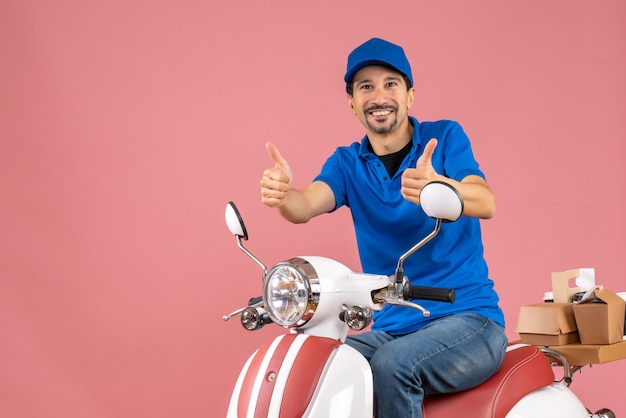 Front view of happy delivery guy wearing hat sitting on scooter making ok gesture on pastel peach background