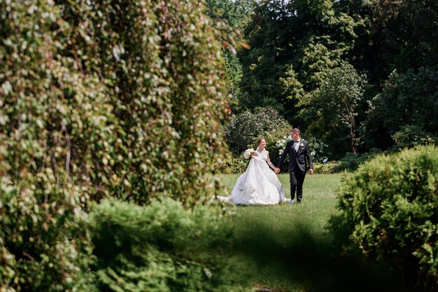 Front view of happy married couple which holding by hands looking to each other while walking together through amazing park during wedding day in summer season