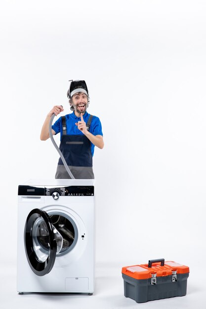 Front view of happy repairman in uniform standing behind washing machine holding pipe on white wall