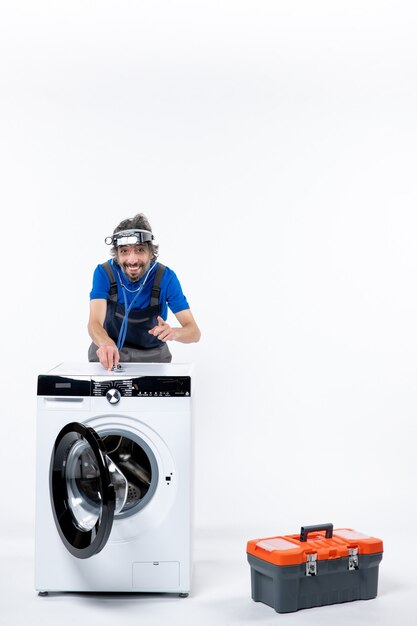 Front view of happy repairman with head lamp putting stethoscope on washer on white wall